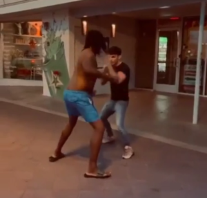 A man in blue trunks initiating an attack with a right hook, while the BJJ practitioner successfully blocks the strike.